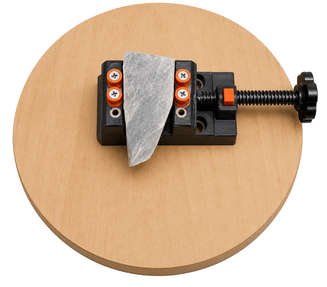 Small drill-press-style vise with soft jaw pads, mounted on a rotating wood disc, holding a flat stone slab